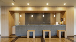 A reception area with a wooden ceiling, a dark brick wall, and three pendant lights. There are three wooden stools in front of a reception desk. - Hotel Route Inn Ube (Ube)
