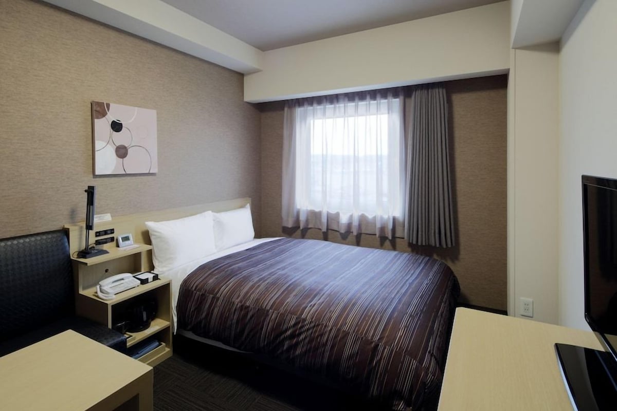 A hotel room with a bed, a desk with a telephone, a flat-screen TV, a bedside table with a lamp, and a window with curtains.