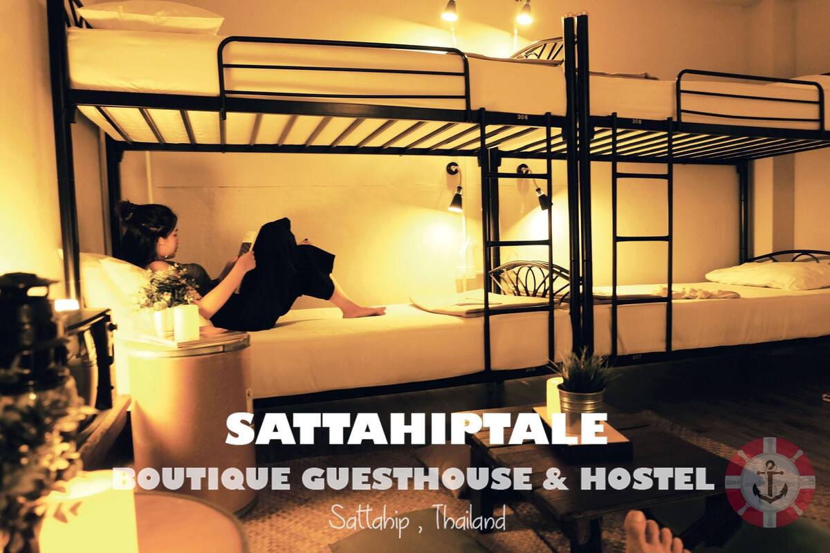A bunk bed room with a person reading a book, a small table with a plant, and a sign indicating it's a boutique guesthouse and hostel in Sattahip, Thailand.