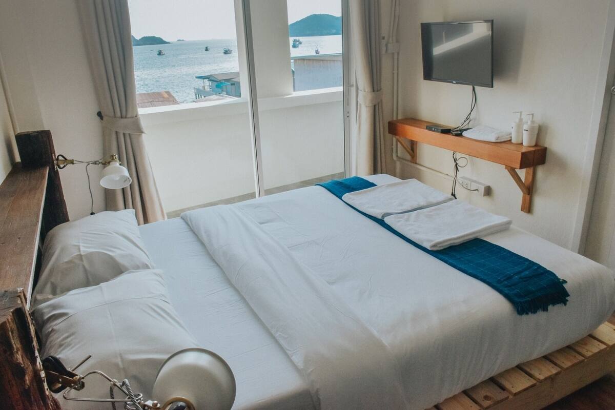 A hotel room with a bed, a TV mounted on the wall, and a view of the sea through the window.