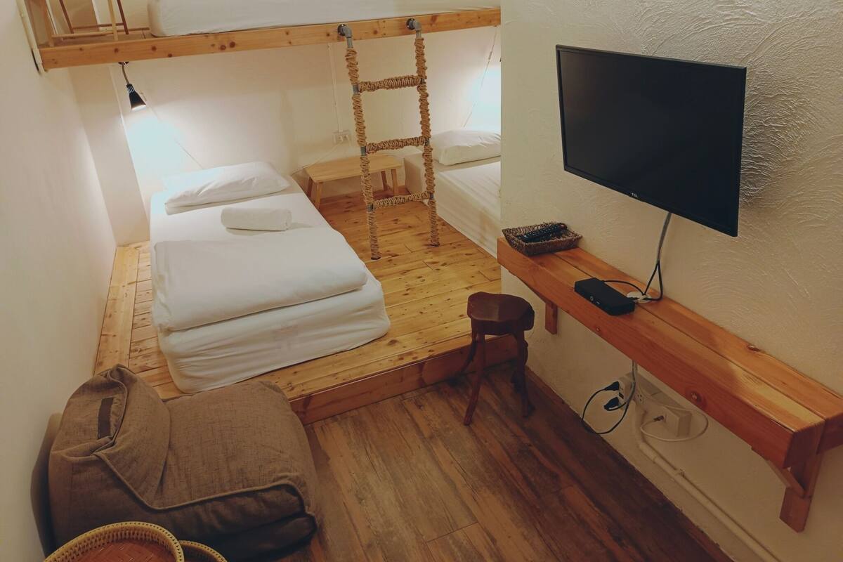 A dorm-style room with bunk beds, a TV, a desk, and a small stool.