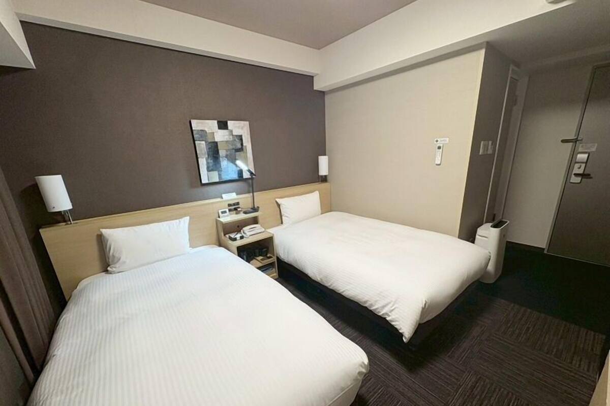 Une chambre d’hôtel avec deux lits, une table de chevet, une lampe, une poubelle et une porte.