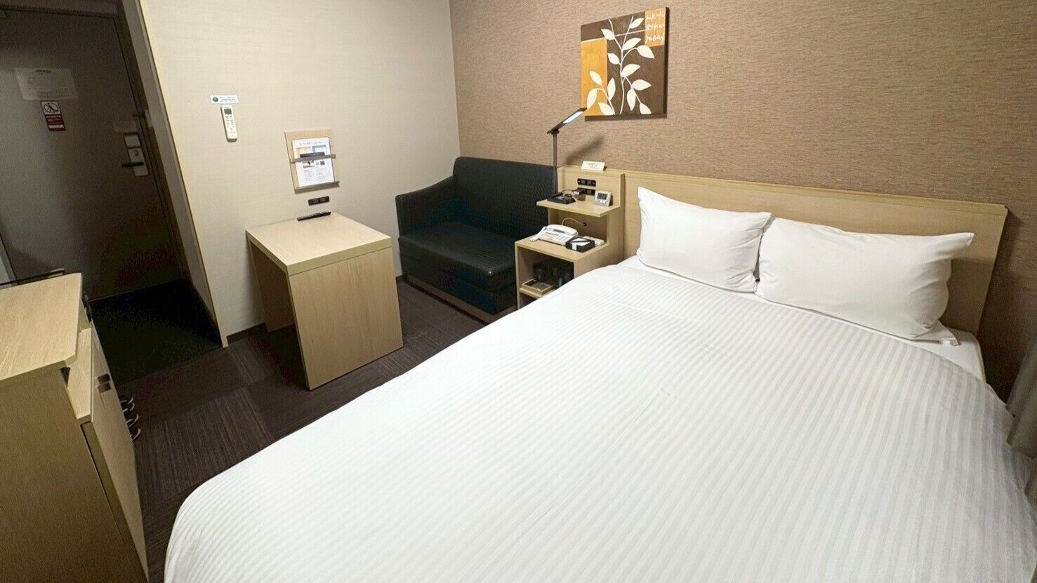 Ein Hotelzimmer mit einem großen Bett, einem Schreibtisch, einem Sessel, einem Nachttisch, einer Lampe und einem an der Wand montierten Telefon.