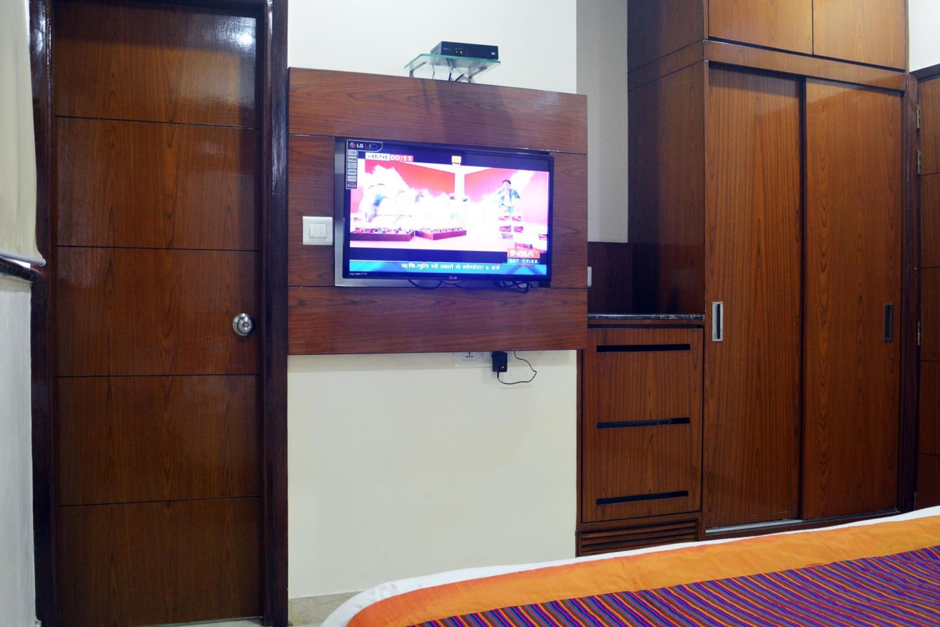 A bedroom with a TV mounted on the wall, wooden wardrobe, and a bed with a colorful bedspread.
