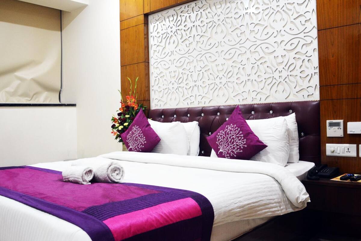 Una cama bien hecha con colcha púrpura y blanca, un cabecero decorativo y una mesita de noche con un teléfono y un control remoto.