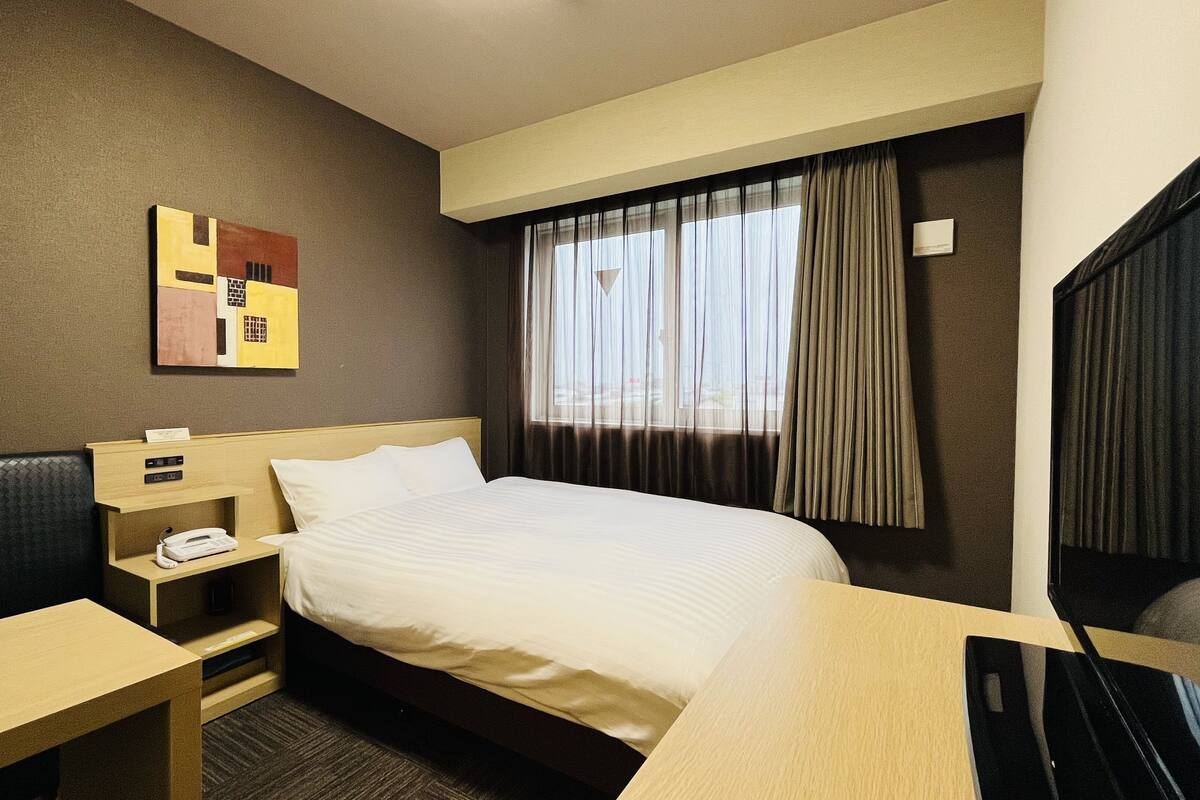 A hotel room with a bed, a desk, a chair, a telephone, and a painting on the wall.