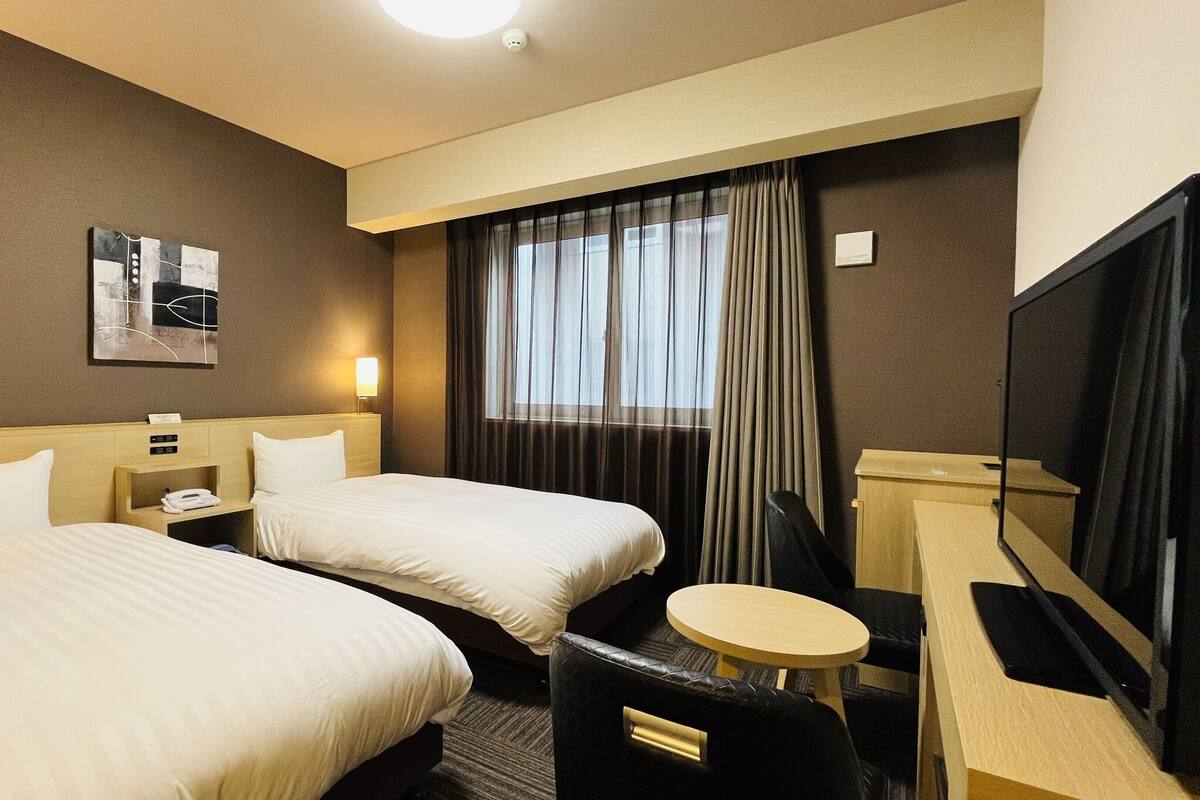 A hotel room with two beds, a desk, a chair, a TV, and a window with curtains.