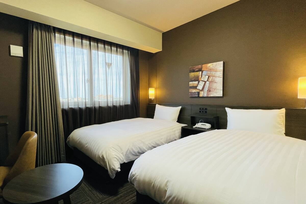 A hotel room with two beds, a desk, a chair, and a window with curtains.