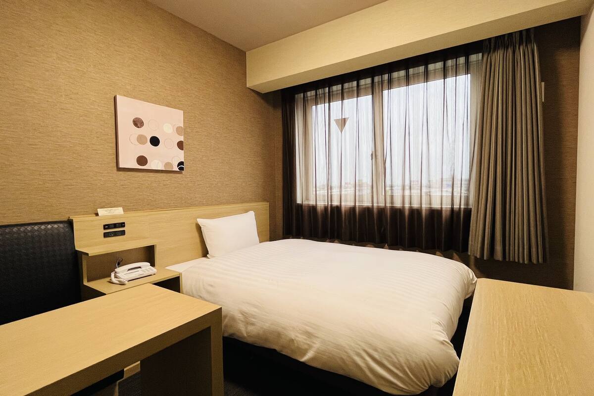 A hotel room with a bed, a desk with a telephone, a television, and a window with curtains.