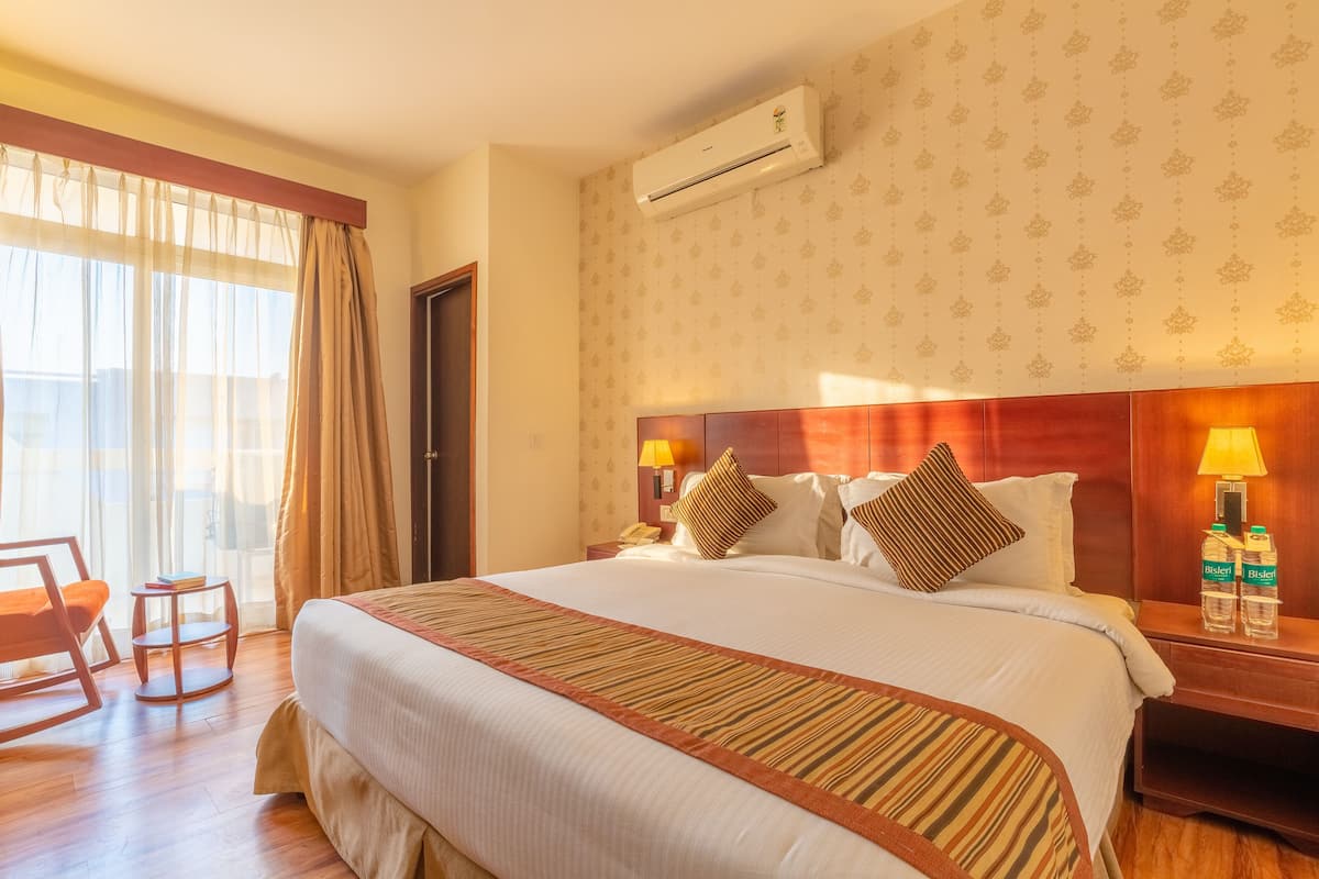 Habitación de hotel con una cama grande, cabecera de madera, mesita de noche con lámpara y ventana con cortinas.