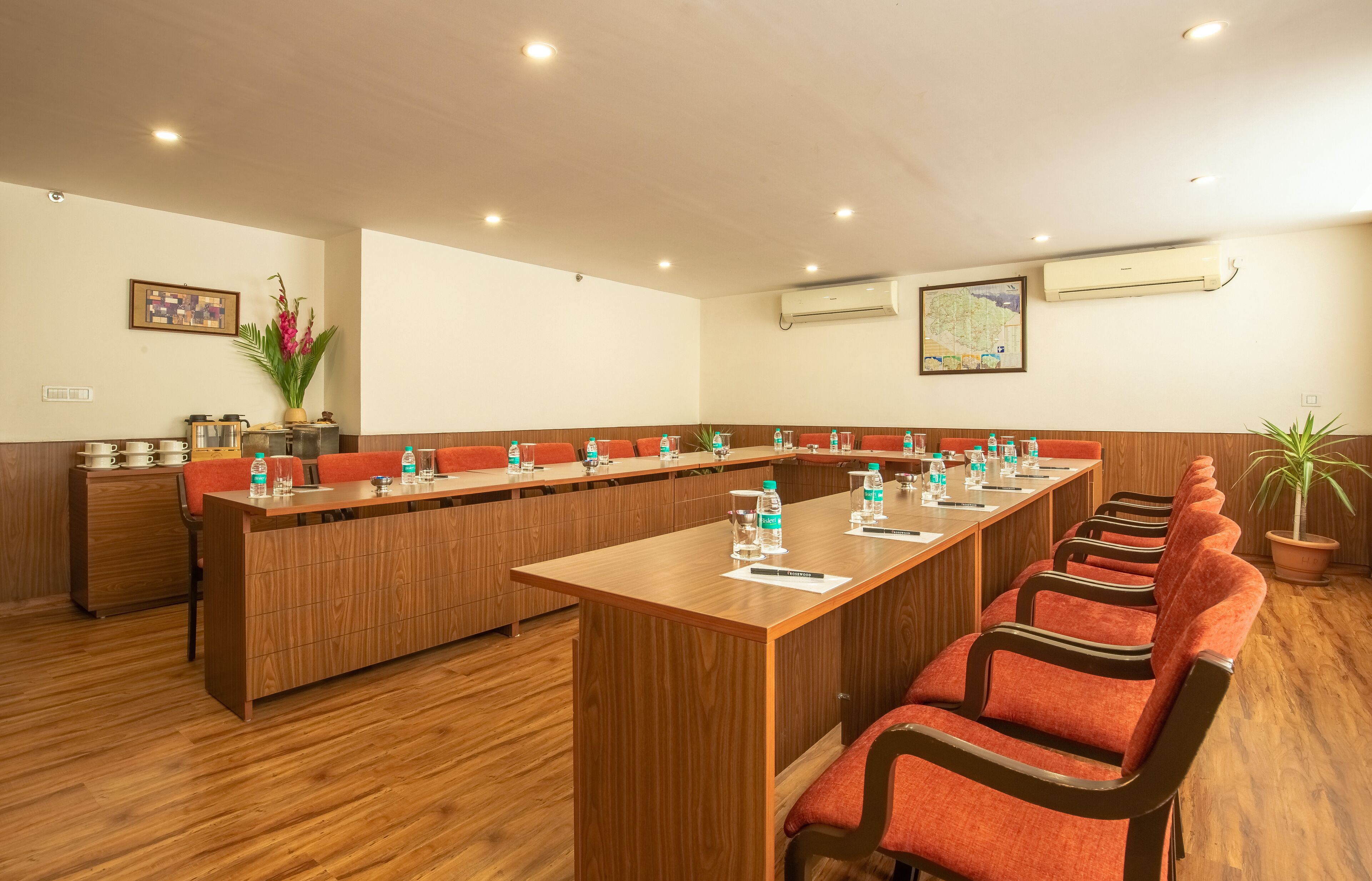 A conference room with a long wooden table, red upholstered chairs, and a wooden cabinet with kitchenware.