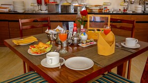 Une table mise pour le petit-déjeuner, avec une nappe pour deux personnes, comprenant des assiettes, des couverts, un verre de jus d’orange, une bouteille d’eau et un vase avec une rose.