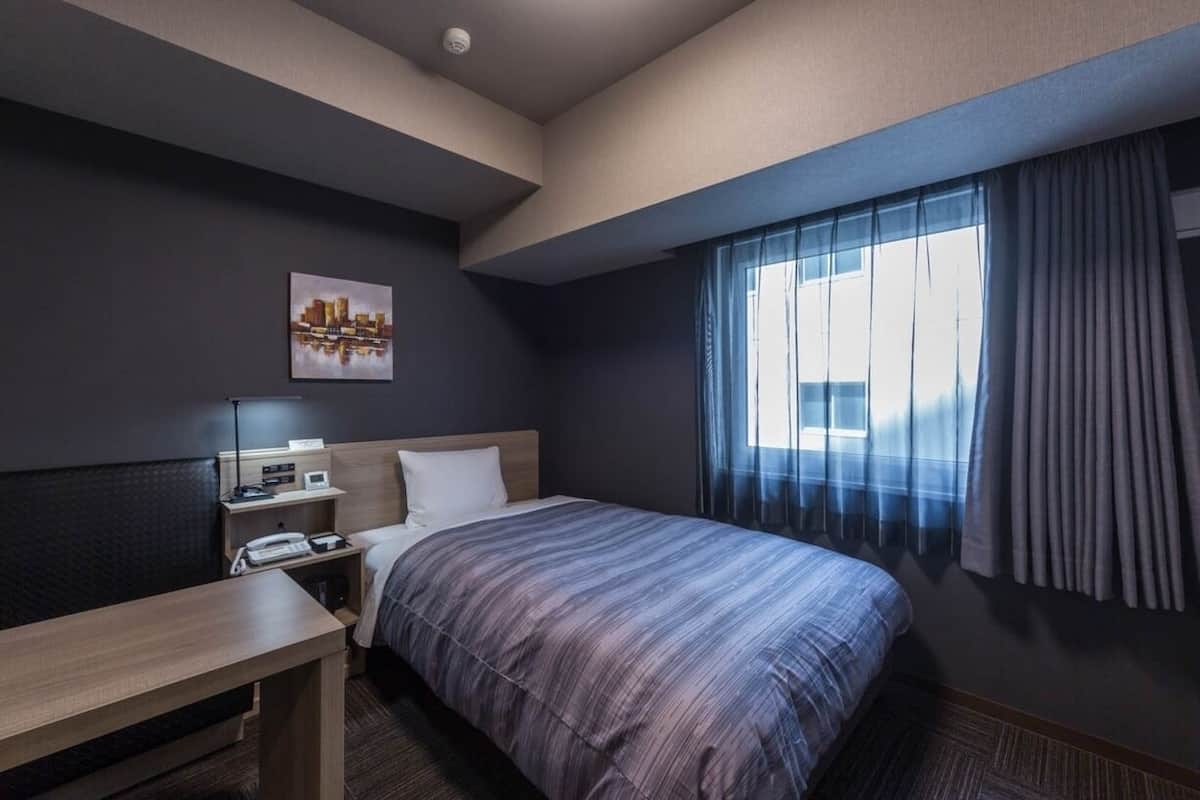 A hotel room with a bed, a desk, a chair, a nightstand, a lamp, a painting, and a window with curtains.