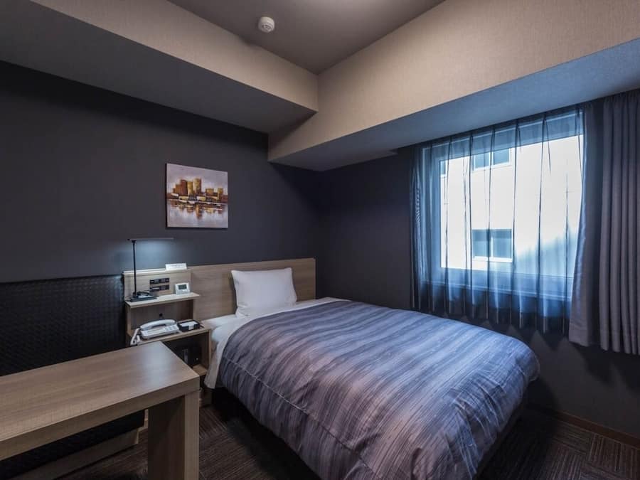 A hotel room with a bed, a desk, a chair, a nightstand, a lamp, a painting, and a window with curtains.