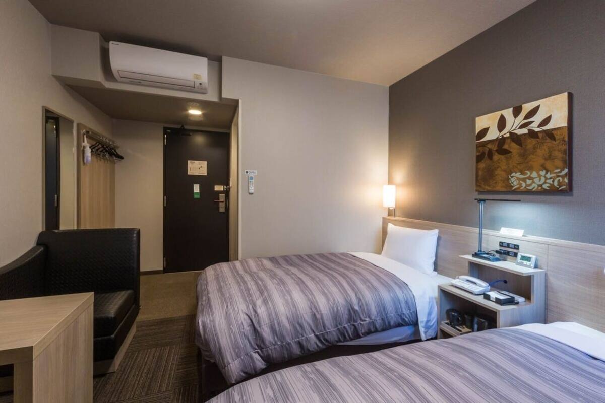 A hotel room with two beds, a desk, a chair, a coat rack, and a wall-mounted artwork.