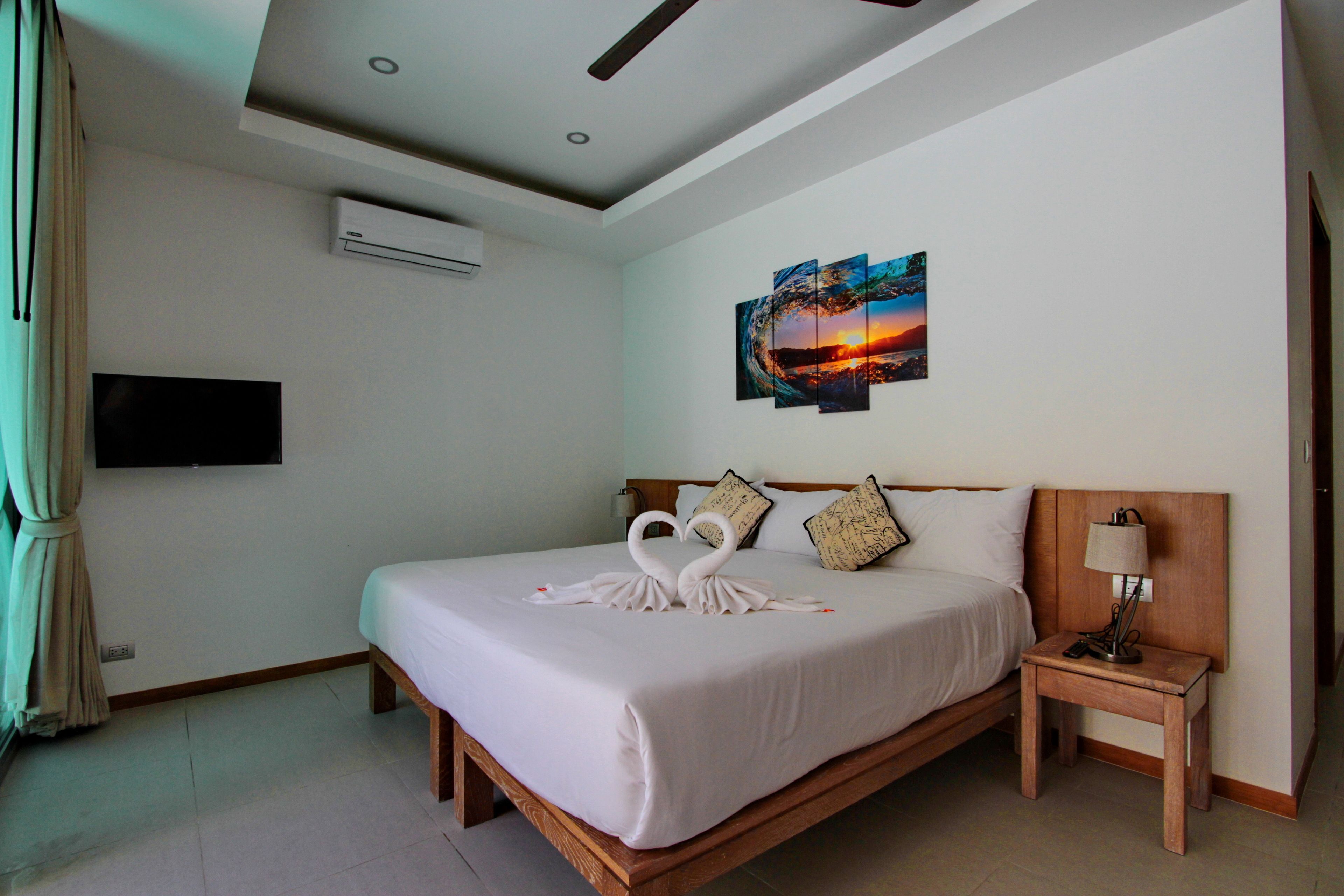A hotel room with a large bed, a flat-screen TV mounted on the wall, a ceiling fan, and a painting of a sunset.