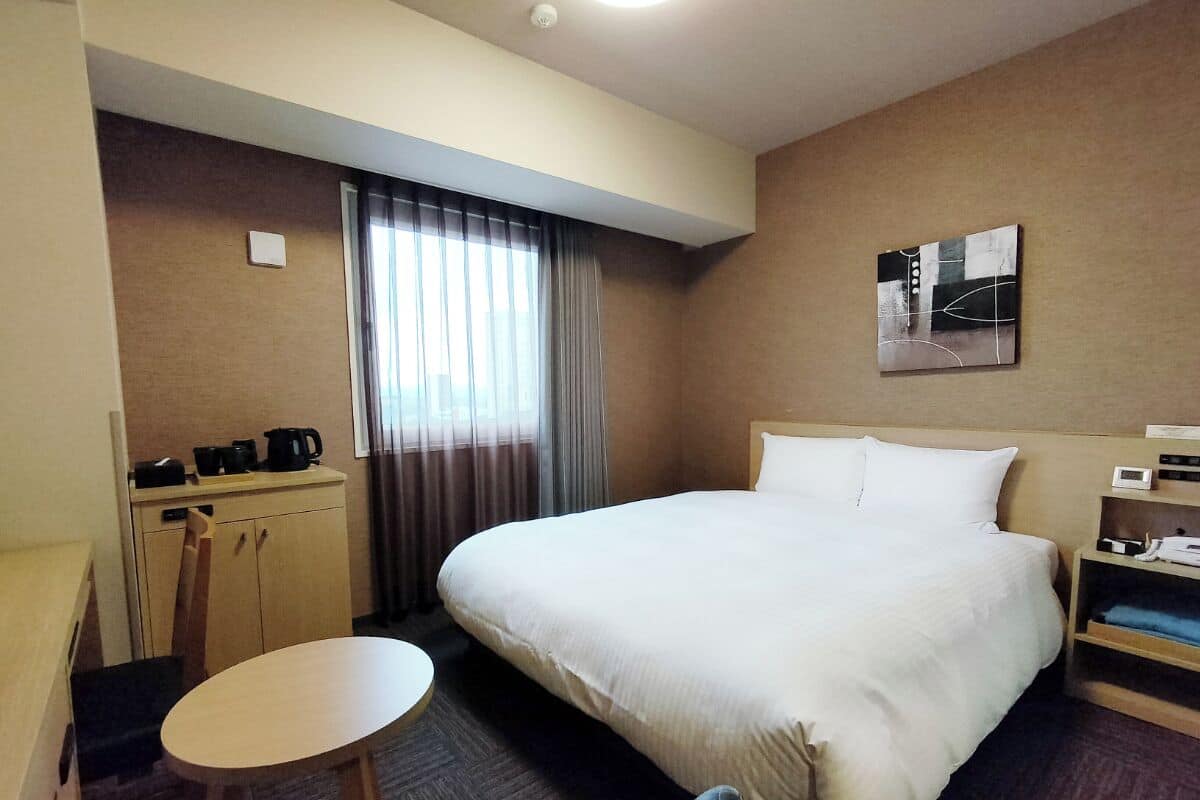 A hotel room with a large bed, a small desk with a chair, a round table, and a window with curtains.