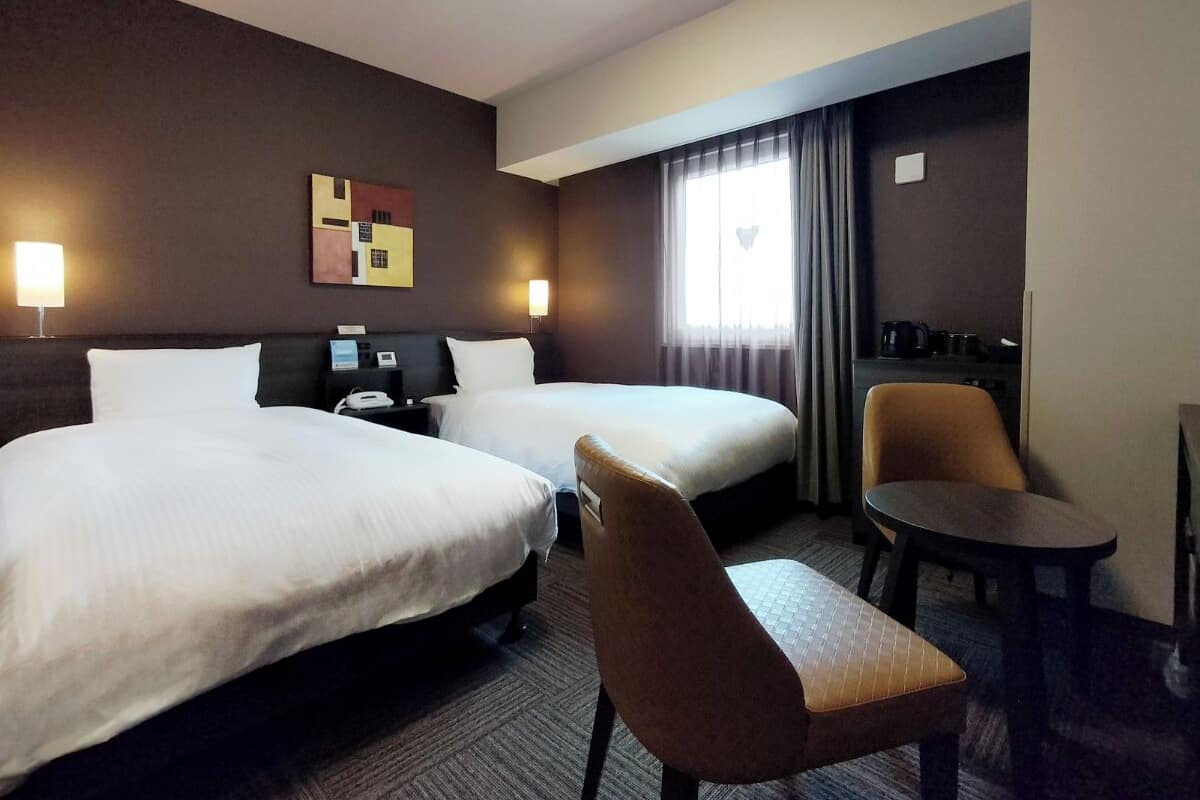 A hotel room with two beds, a desk, a chair, and a small table.