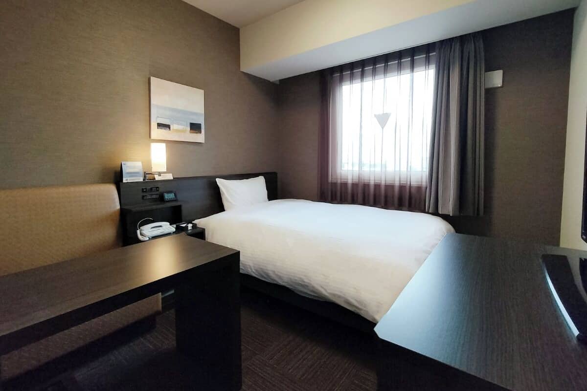 A hotel room with a bed, a desk, a nightstand, a lamp, and a window with curtains.