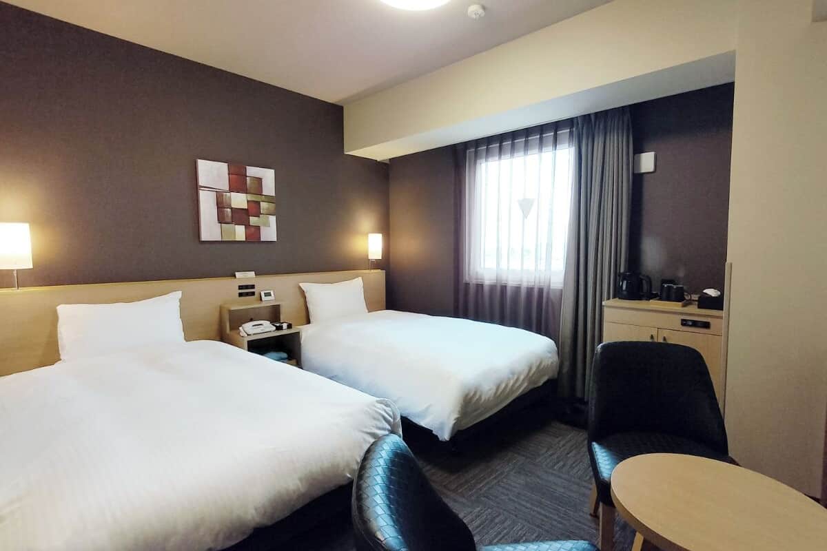 A hotel room with two beds, a desk, a chair, a small table, and a window with curtains.
