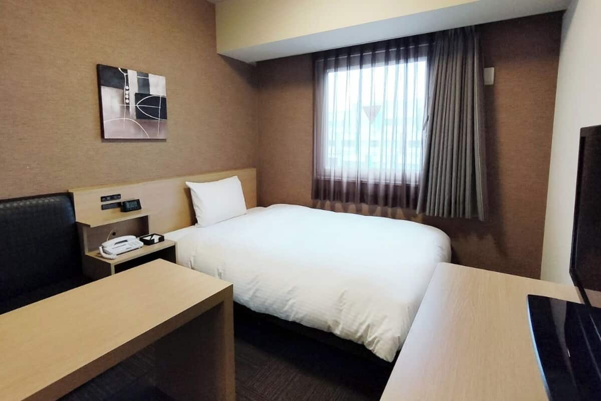A hotel room with a bed, a desk, a chair, a TV, a clock, and a window with curtains.