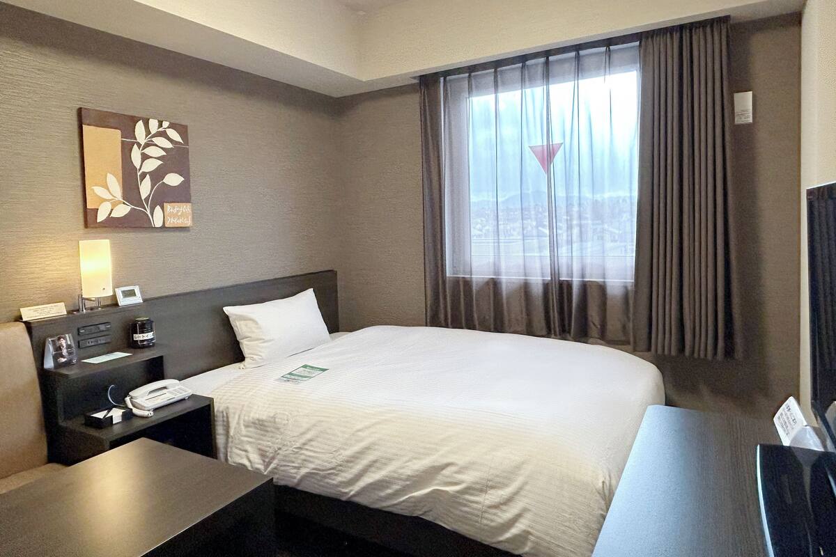 Ein Hotelzimmer mit Bett, Schreibtisch, Telefon, Lampe und einem fenster mit Vorhängen.