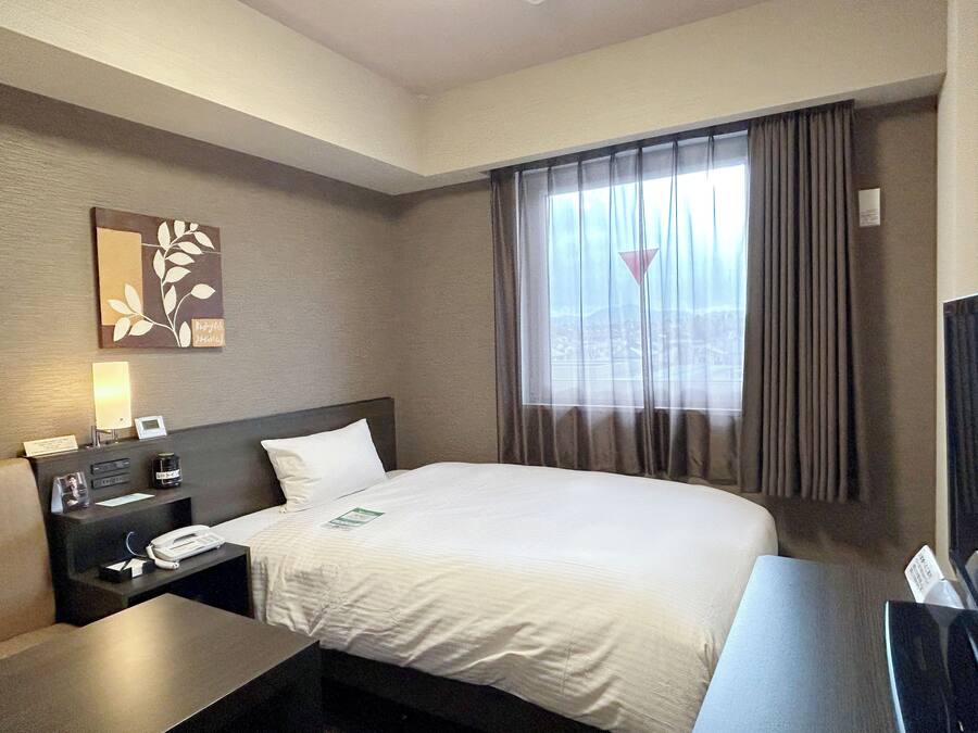 A hotel room with a bed, a desk, a telephone, a lamp, and a window with curtains.
