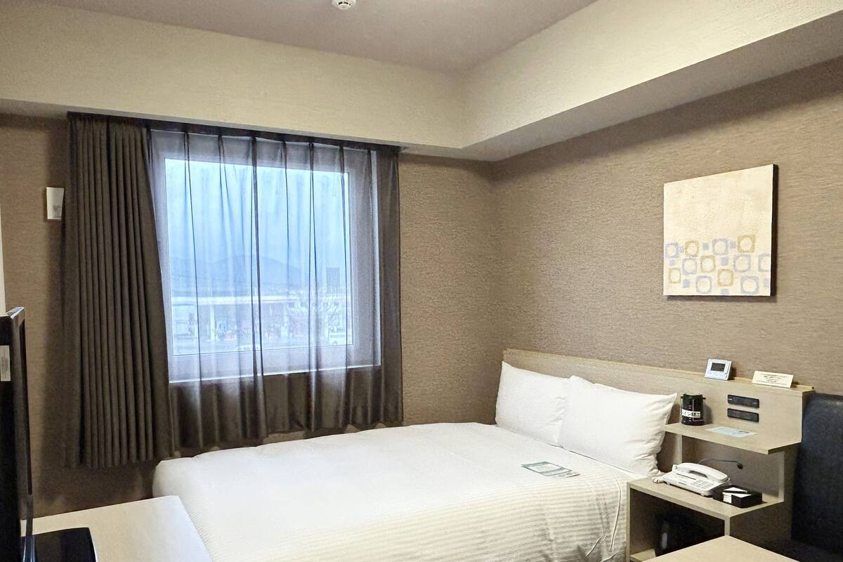Ein Hotelzimmer mit einem großen Bett, einem Nachttisch mit Telefon und Lampe, einem Fenster mit Vorhängen und einem an der Wand hängenden Kunstwerk.