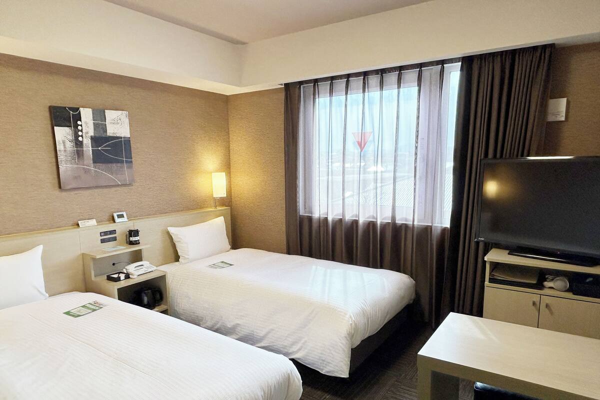 Ein Hotelzimmer mit zwei Betten, einem Schreibtisch, einem Fernseher und einem Fenster mit Vorhängen.