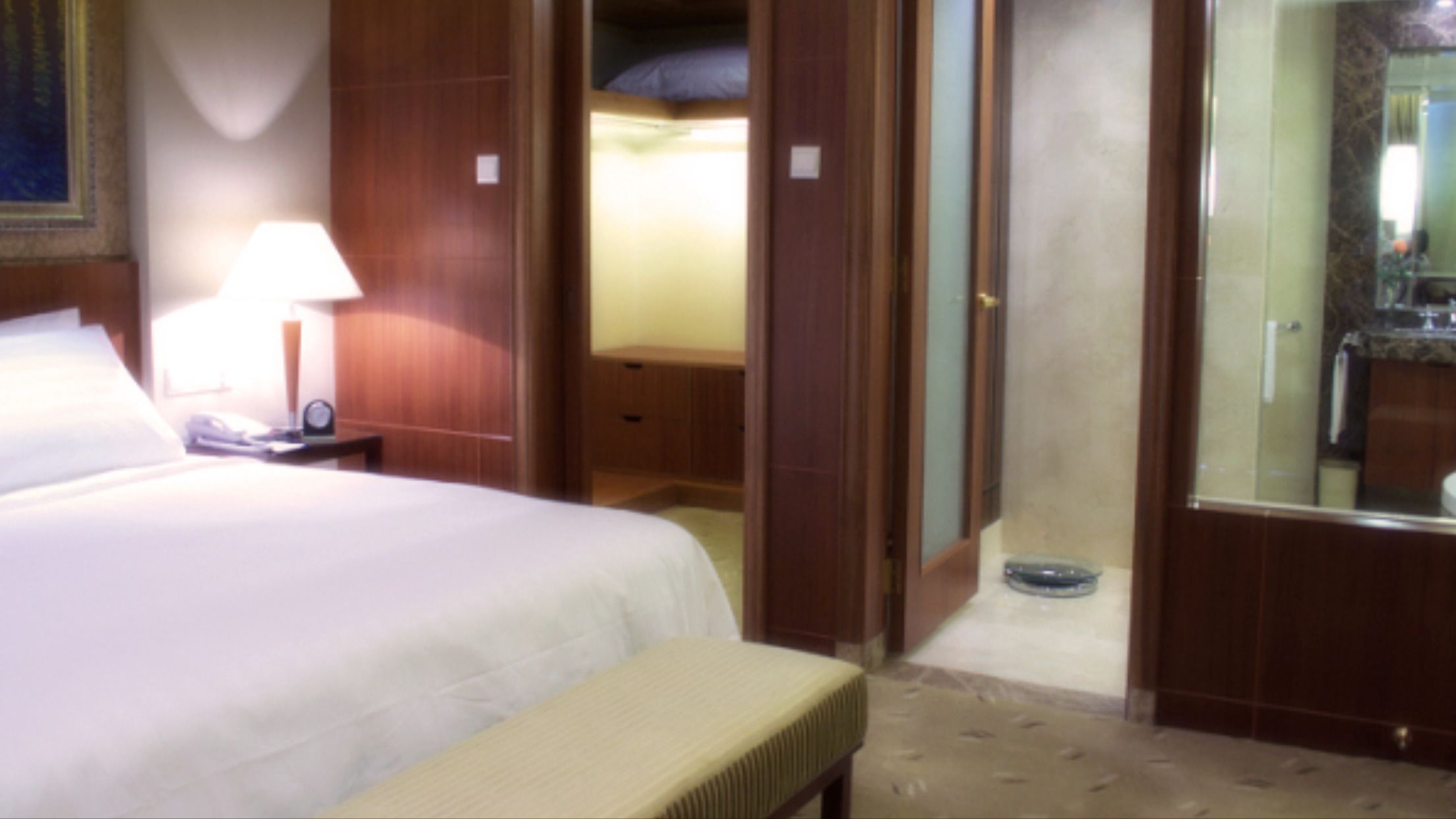A hotel room with a bed, a bedside table with a lamp, a bench, a wardrobe, and a bathroom with a mirror and sink.