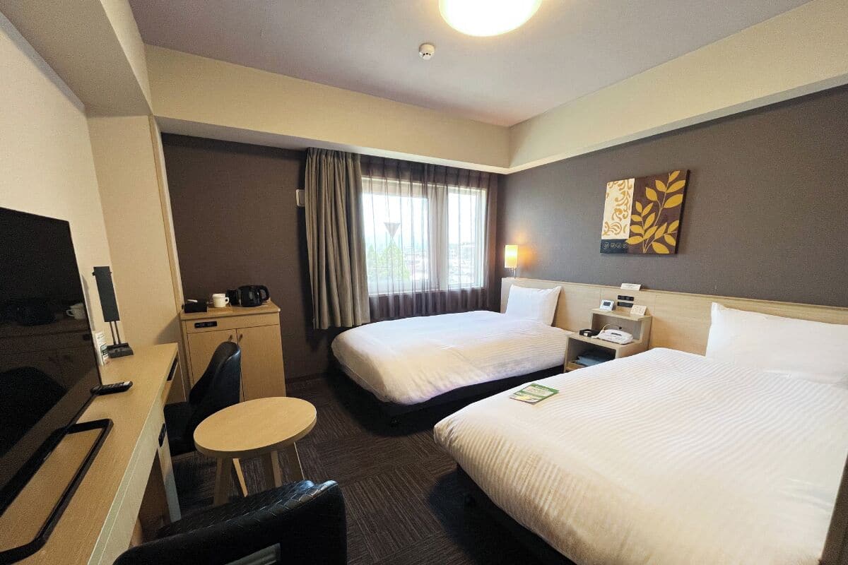 A hotel room with two beds, a desk with a chair, a small table, a TV, and a window with curtains.