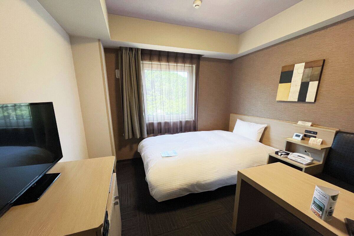 A hotel room with a bed, a desk, a TV, a nightstand, and a window with curtains.