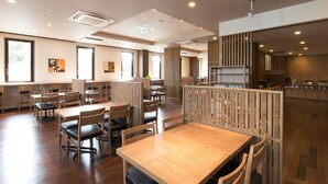 Un restaurant moderne avec des tables et des chaises en bois, de grandes fenêtres et un espace cuisine visible à travers une cloison.