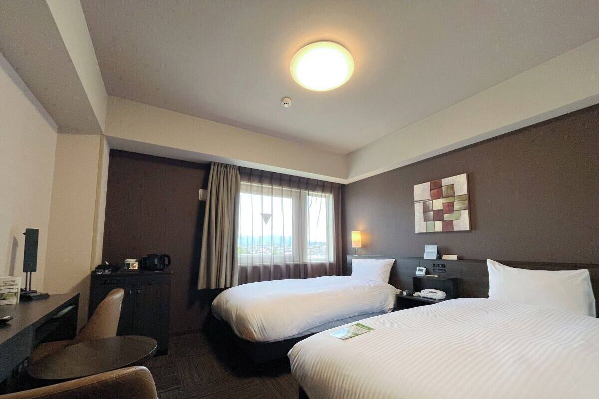 A hotel room with two beds, a desk with a chair, a small table, a lamp, a nightstand, and a window with curtains.