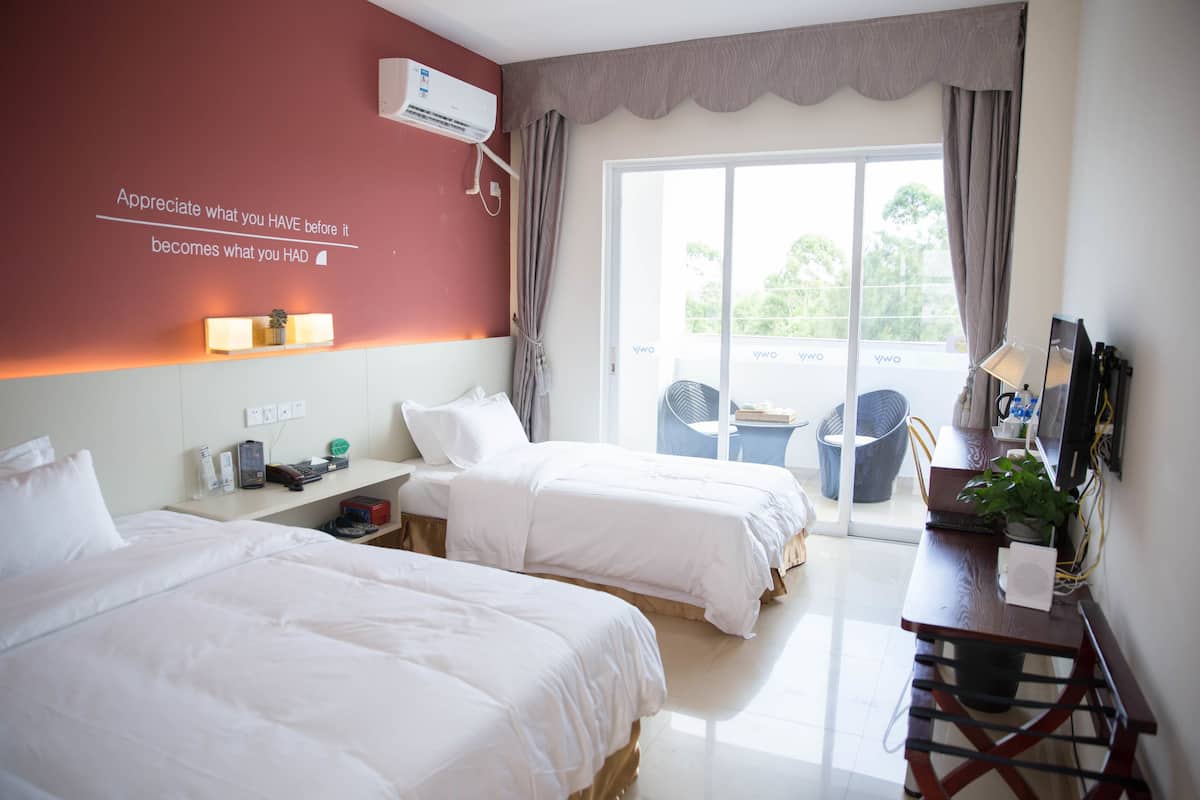 A hotel room with two beds, a desk, a chair, a potted plant, and a large window with curtains.