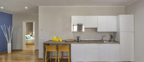 Una cocina moderna con armarios blancos, un lavamanos de acero inoxidable y un horno empotrado. Hay dos sillas de madera junto a una mesita pequeña, y una planta en maceta sobre la encimera.