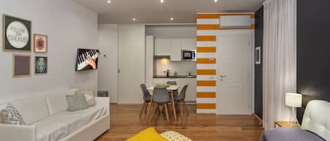 A modern hotel room with a sofa, a bed, a dining area, and a kitchen in the background.