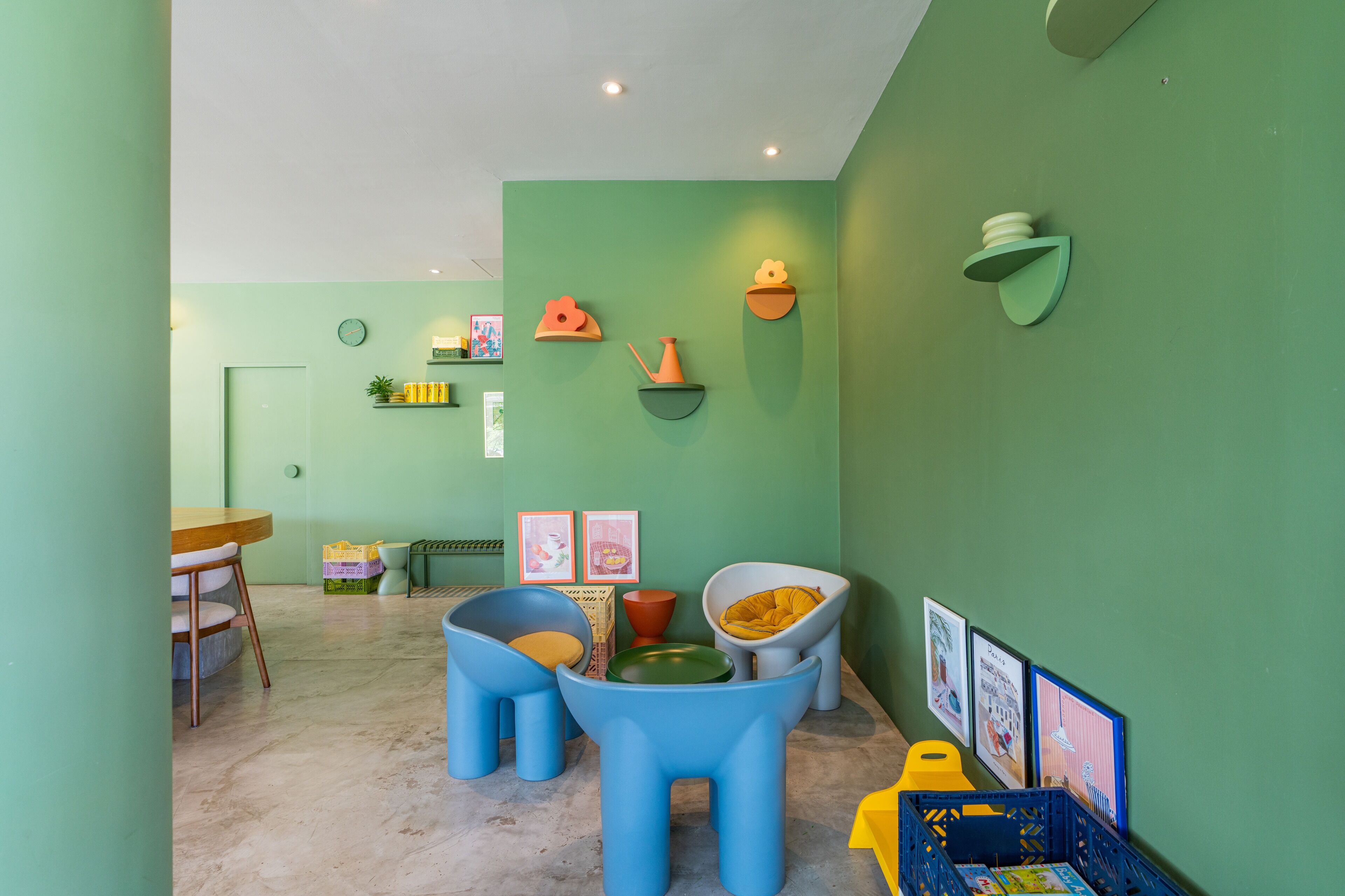 A room with green walls, a wooden table, and blue and yellow chairs. There are wall-mounted shelves with various items and framed pictures.