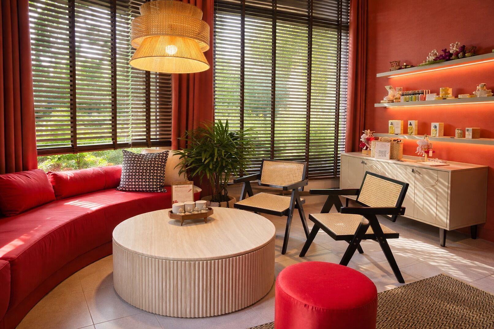 A modern living room with a red sofa, a round coffee table, wicker chairs, a cabinet with shelves, and large windows with blinds.