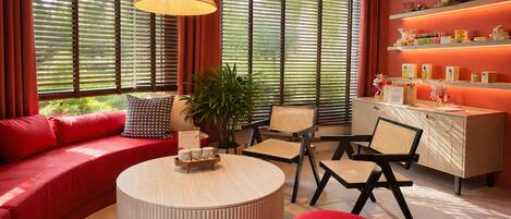 A modern living room with a red sofa, a round coffee table, wicker chairs, a cabinet with shelves, and large windows with blinds.