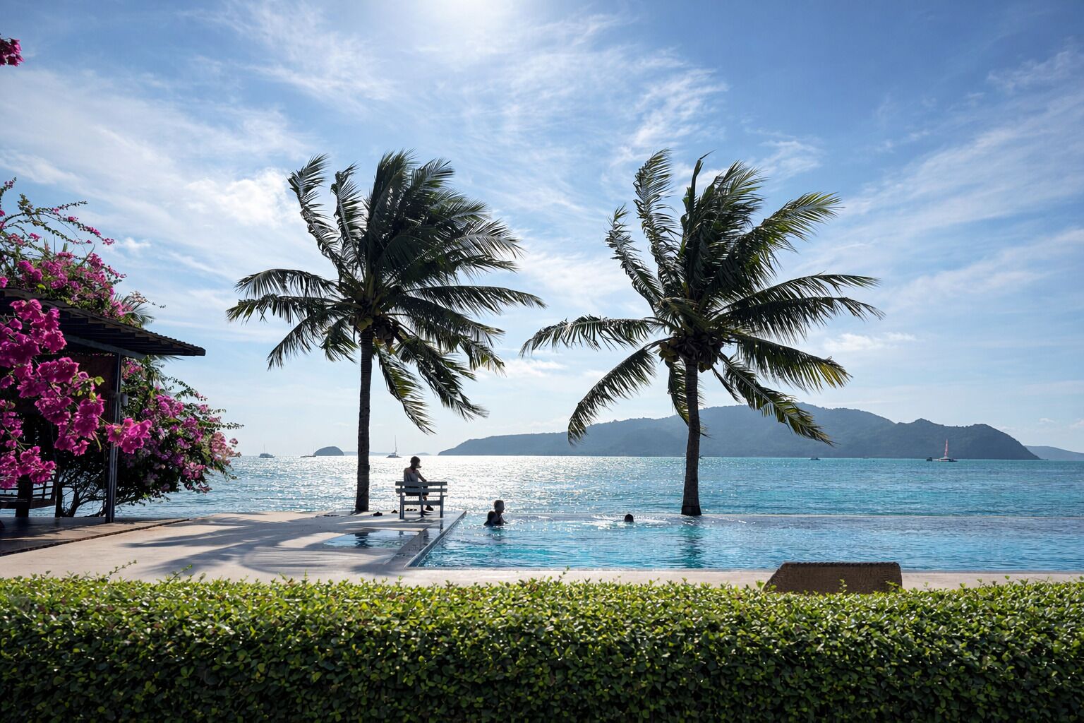 Un espace aménagé au bord de la piscine, avec deux palmiers, un banc et une vue sur la mer.