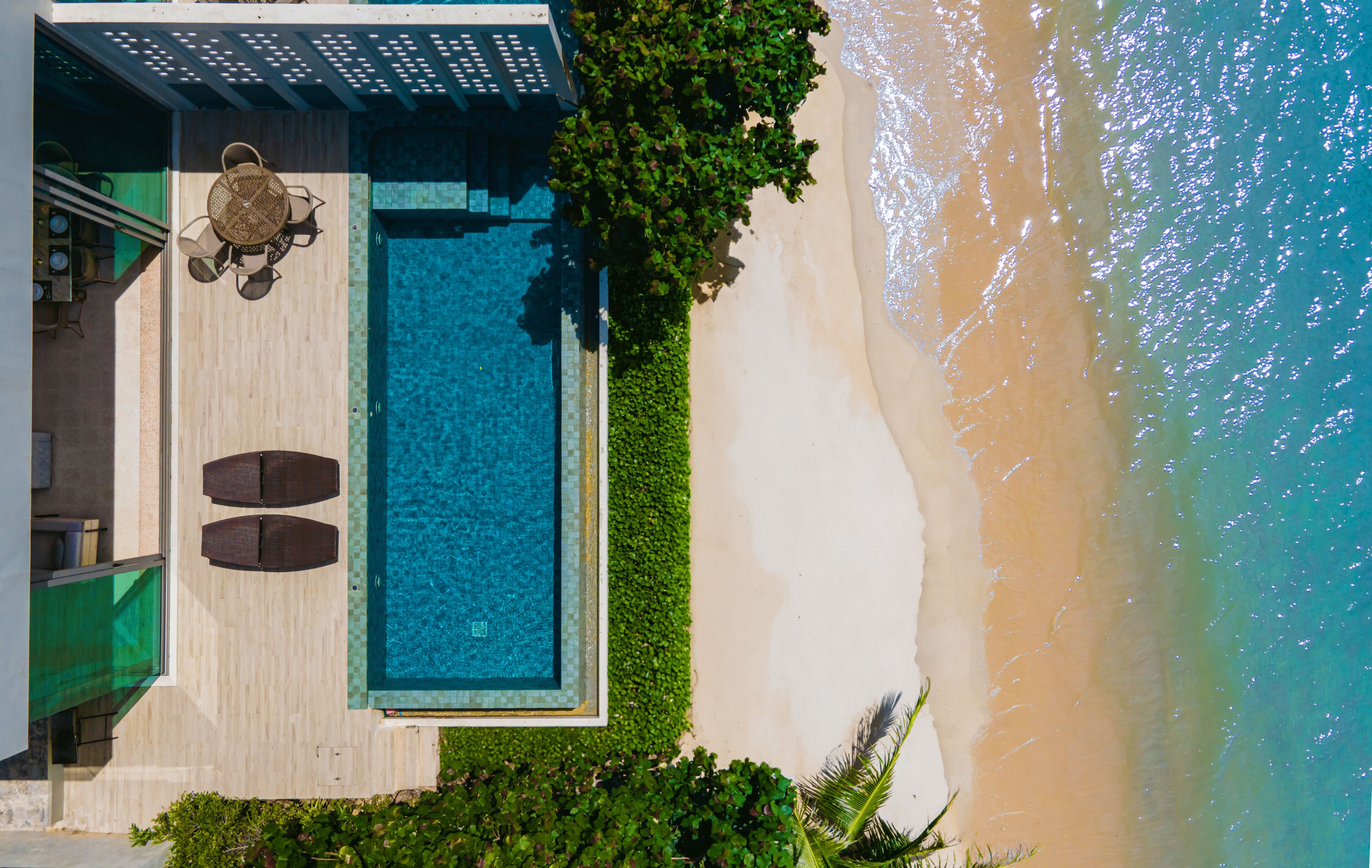 Une vue aérienne d’une piscine située à côté d’une plage, avec un espace patio aménagé avec du mobilier d’extérieur.