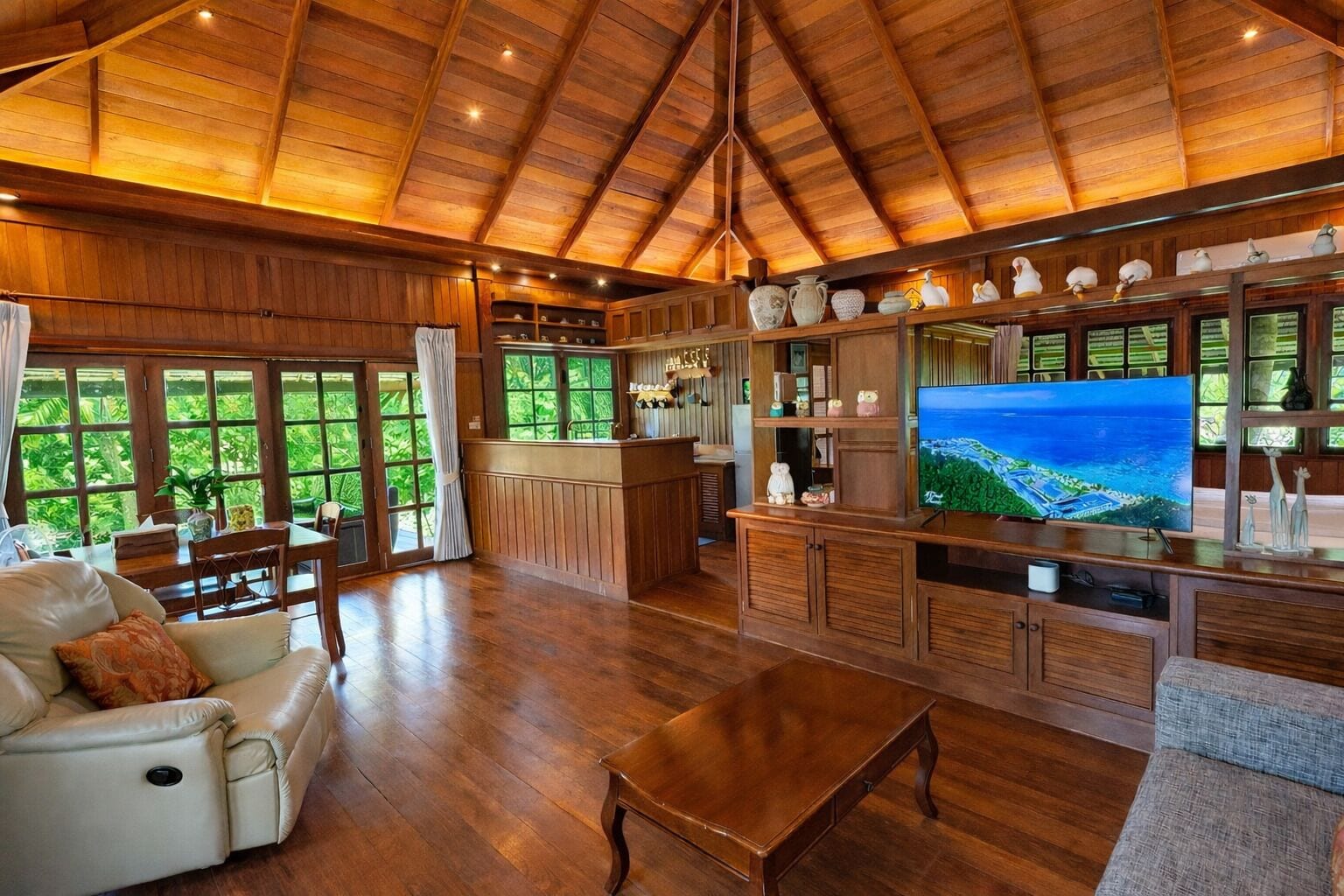 A spacious living area with a wooden ceiling, a large TV, and a view of greenery through the windows.