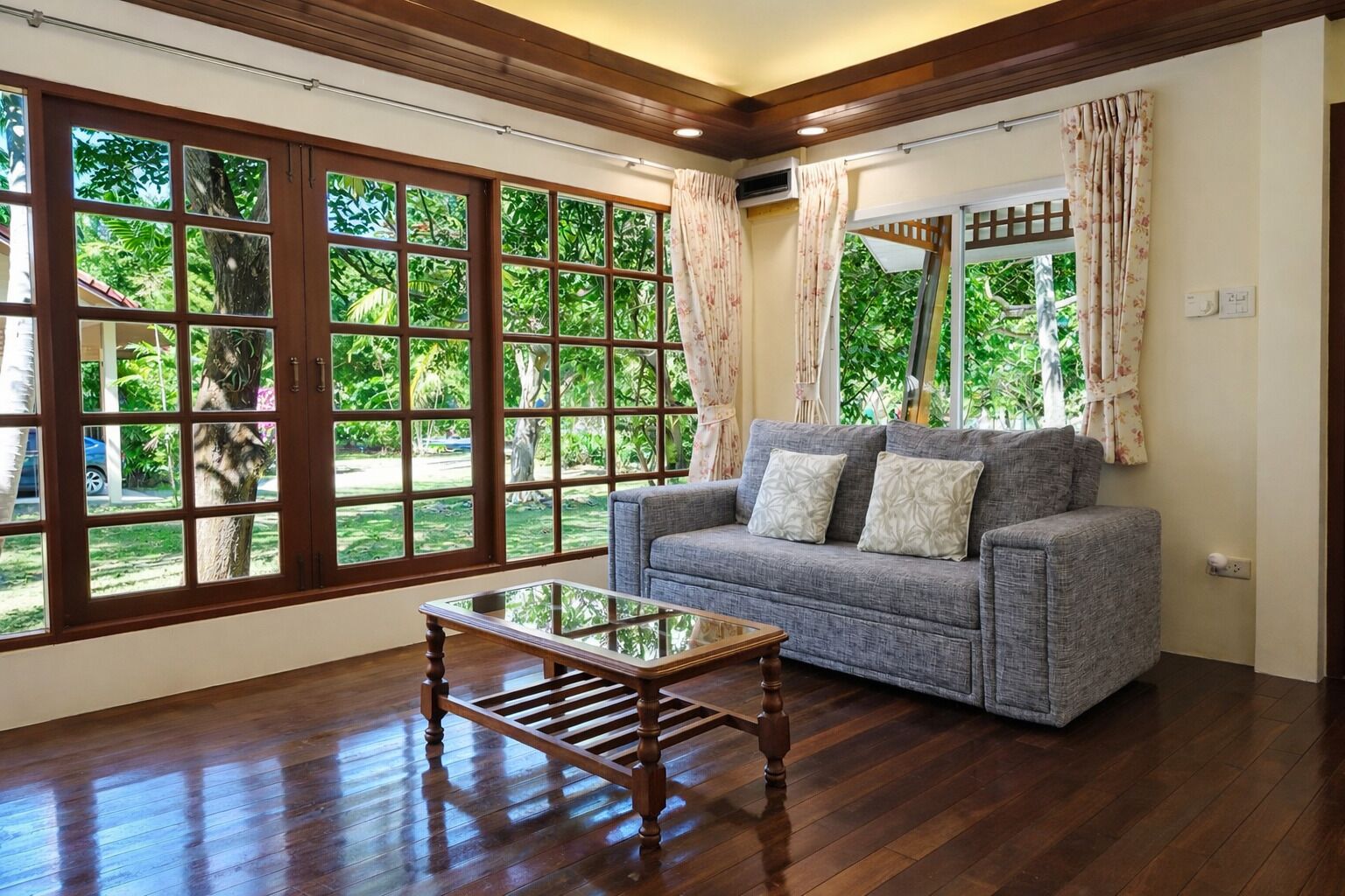 A room with a grey sofa, a glass coffee table, wooden flooring, large windows with curtains, and an air conditioning unit.