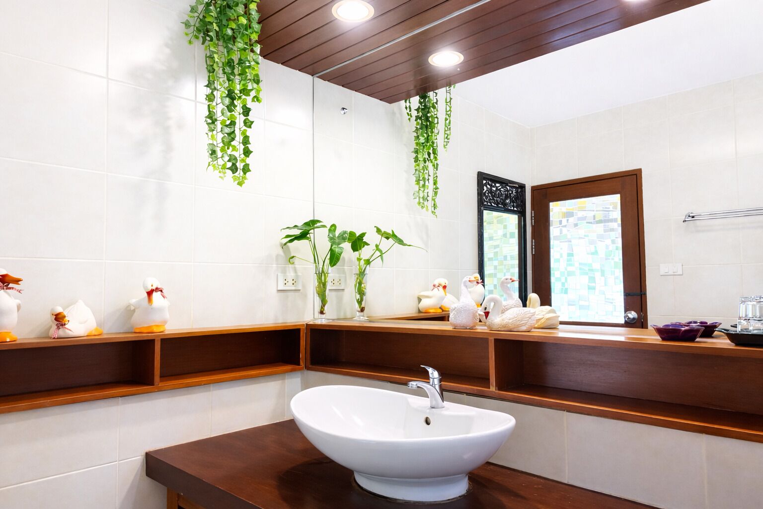 Ein Badezimmer mit einem weißen Waschbecken, Holzregalen, hängenden Pflanzen und einem Spiegel mit dekorativem Rahmen.
