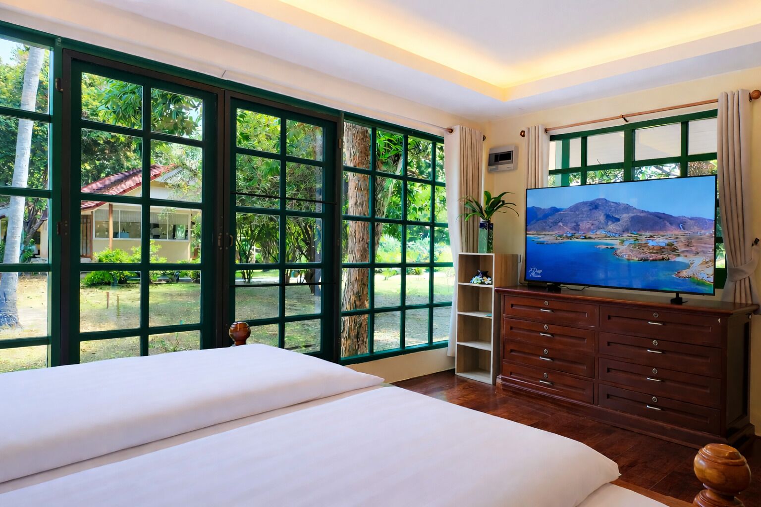 A spacious bedroom with a large bed, a TV, and a view of a lush garden through large windows.