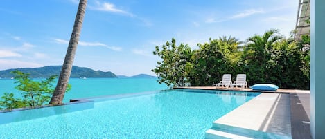 An infinity pool with clear blue water, white lounge chairs, and a view of the sea and distant mountains.