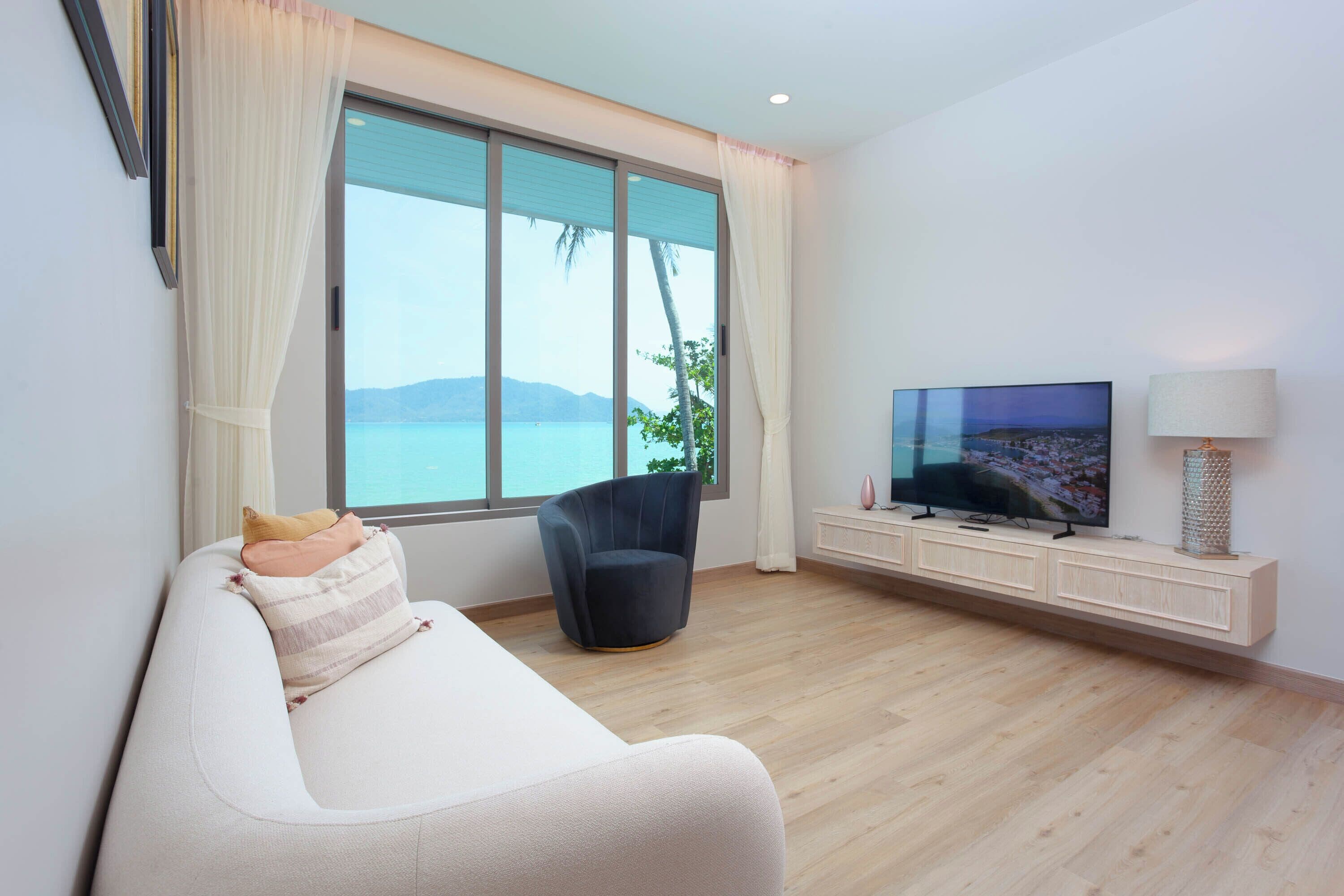 Ein modernes Wohnzimmer mit einer Couch, einem schwarzen Sessel, einem Fernseher und einem großen Fenster mit Meerblick.