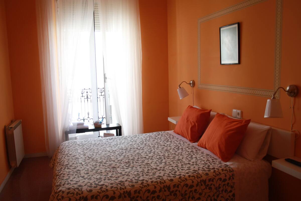 A hotel room with a bed, two bedside lamps, a nightstand, and a window with sheer curtains.