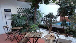 Un patio al aire libre con mesas y sillas de madera, rodeado de plantas y una piscina.